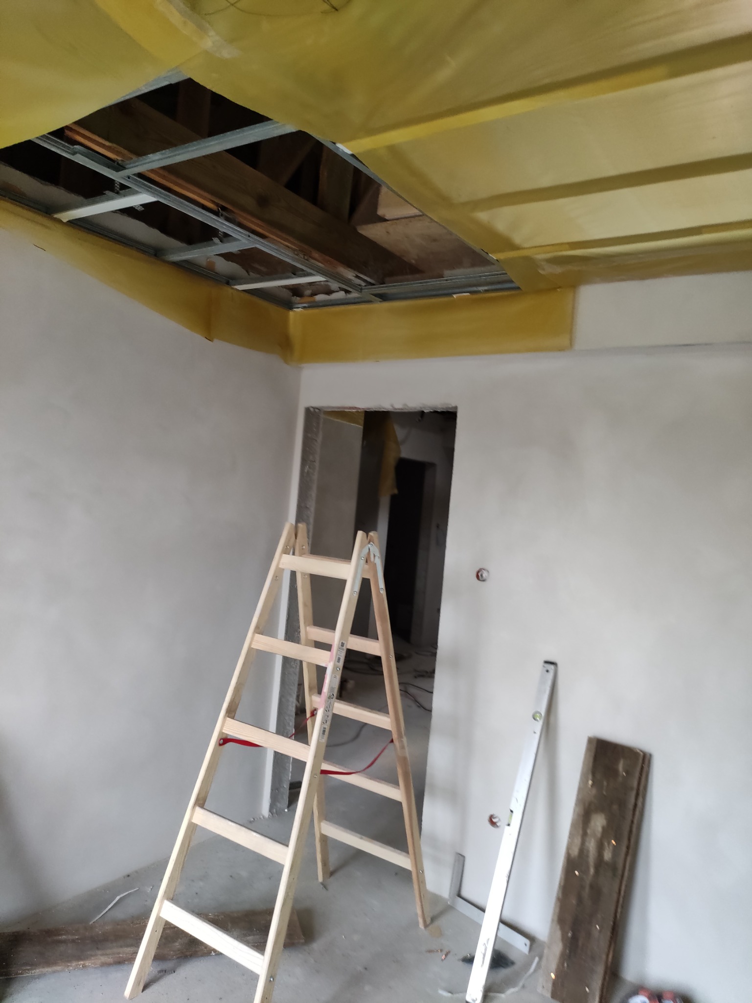 Wnętrze w trakcie montażu sufitu podwieszanego z widoczną metalową konstrukcją i częściowo zamontowaną żółtą izolacją, drewniana drabina oparta o ścianę, poziomica i deska przy ścianie.