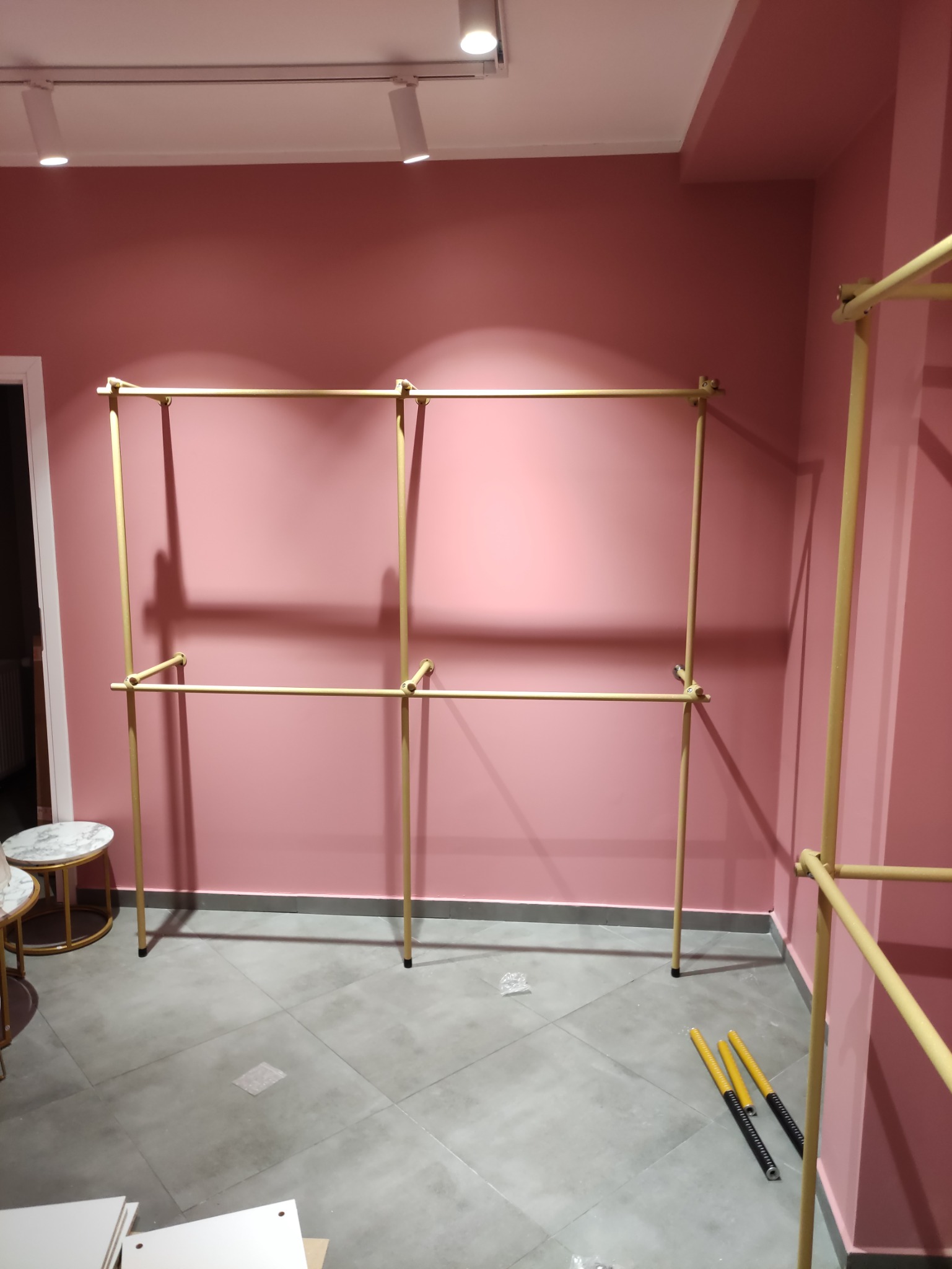 Metalowy stelaż na ubrania w trakcie montażu, widoczne elementy konstrukcyjne i narzędzia na szarych płytkach podłogowych, różowe ściany w tle, oświetlenie sufitowe.