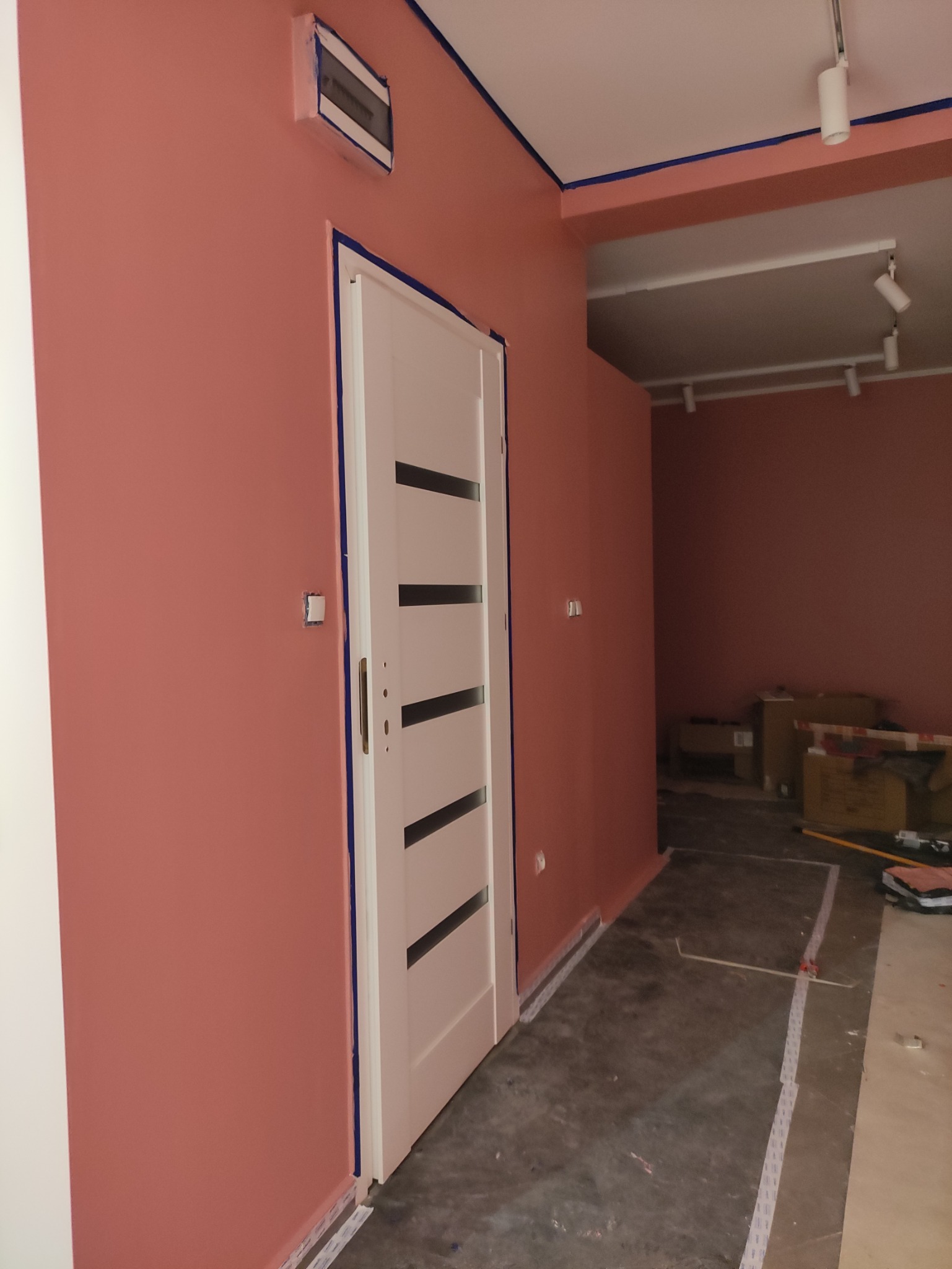 Ściana w odcieniach różu, świeżo pomalowana w mieszkaniu w Białej Podlaskiej, zabezpieczona taśmą malarską przy suficie, podłodze i ościeżnicy białych drzwi z czarnymi paskami. Na ścianie widoczna...