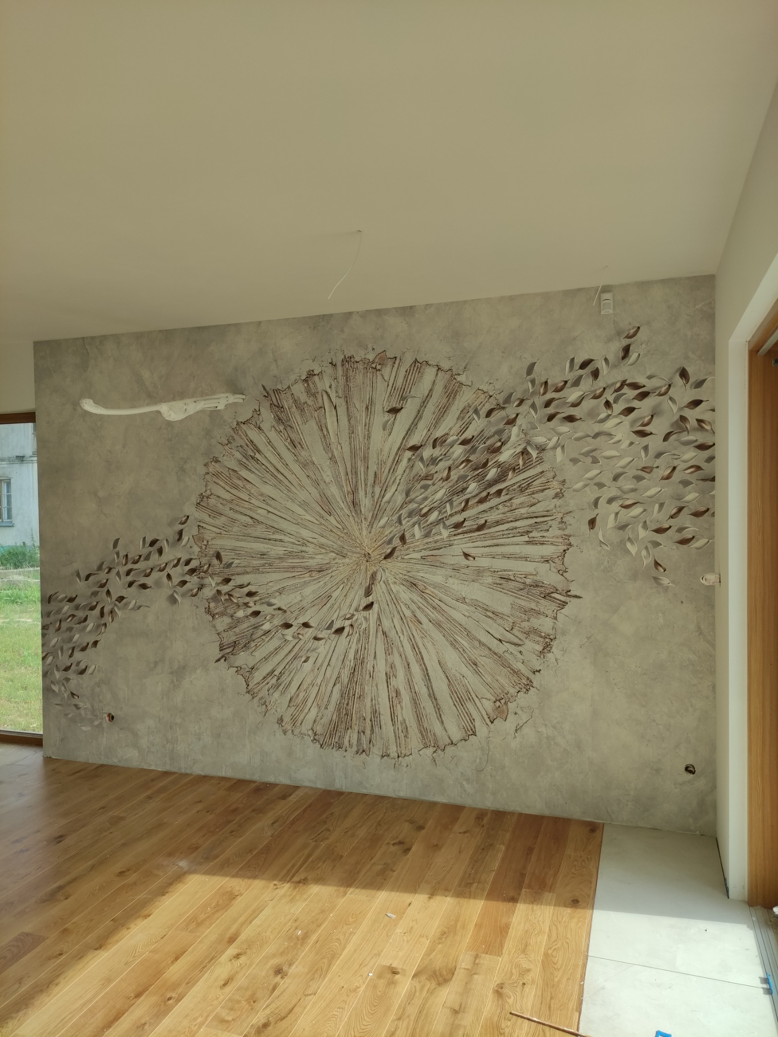 Dekoracyjna ściana z efektem słońca i liści w przestronnym wnętrzu, widoczna drewniana podłoga i fragment okna.