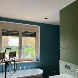 MARK-MAL Marek Leśniak - Świeżo pomalowana łazienka z wanną w mozaikę, umywalką na szafce i oknem. Widoczne oświetlenie robocze i wiadro z gruntem, co sugeruje etap wykończeniowy.