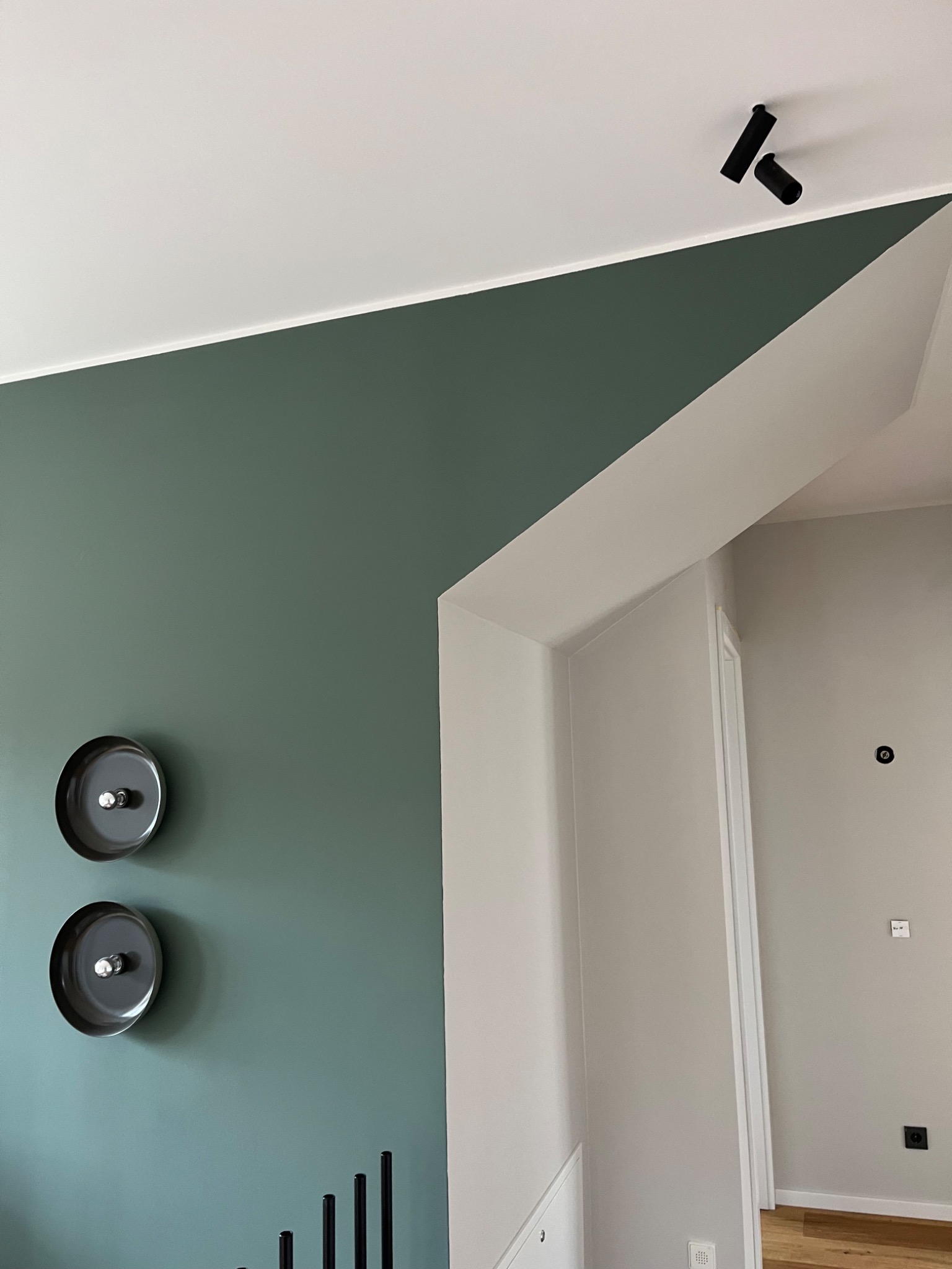 Ściana pomalowana na zielono z dekoracyjnymi talerzami i czarnymi świecznikami, biały sufit z reflektorami. Widok na sąsiednie pomieszczenie przez białe przejście.