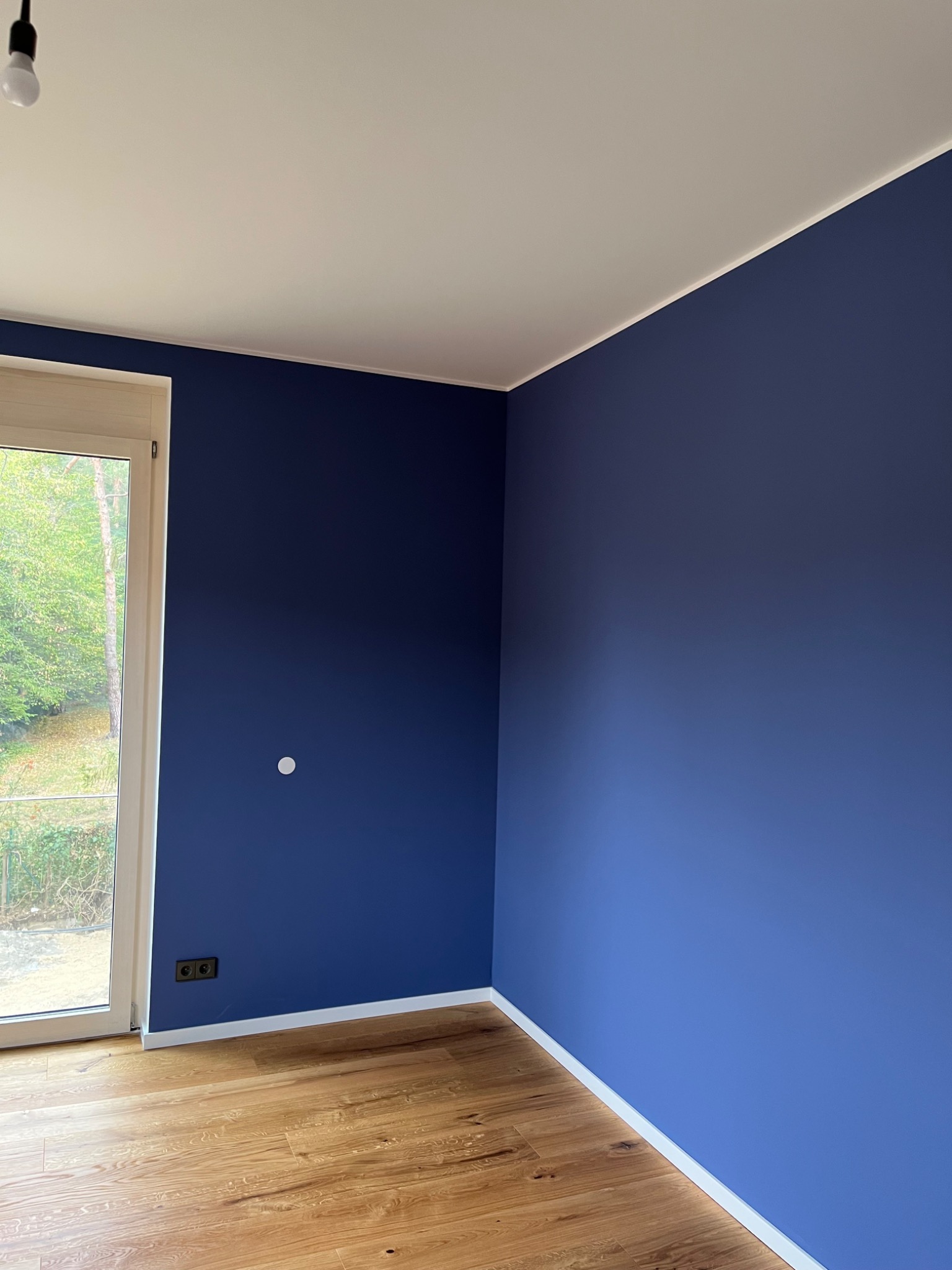 Pomieszczenie po malowaniu ścian na intensywny niebieski kolor. Widoczne drewniane panele podłogowe, biały sufit i okno z widokiem na zieleń. Kontrastowe zestawienie kolorów.