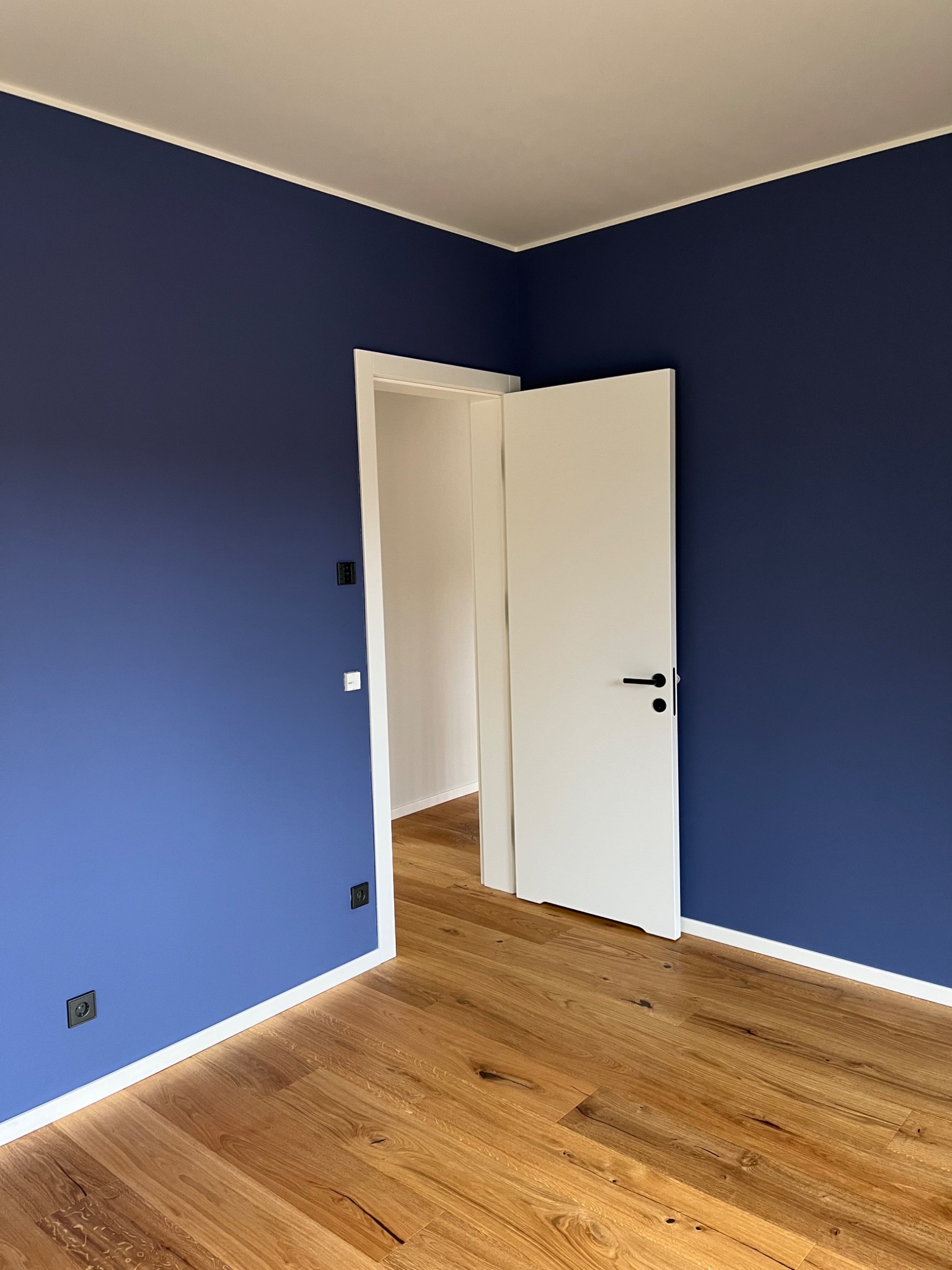 Pokój po malowaniu: ściany w odcieniu granatu, drewniana podłoga, białe listwy przypodłogowe i drzwi. Widoczne czarne gniazdka elektryczne.