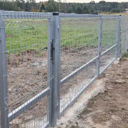 Nowo postawione, srebrne ogrodzenie panelowe z bramką, połączone z siatką, teren wyrównany, widoczne ślady po pracy ziemnej, tło z drzewami i drogą.