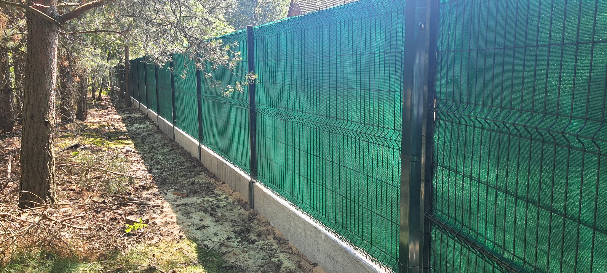 Długi fragment zielonej siatki maskującej na ogrodzeniu panelowym z podmurówką betonową, widok z boku wzdłuż linii ogrodzenia na tle leśnego krajobrazu.