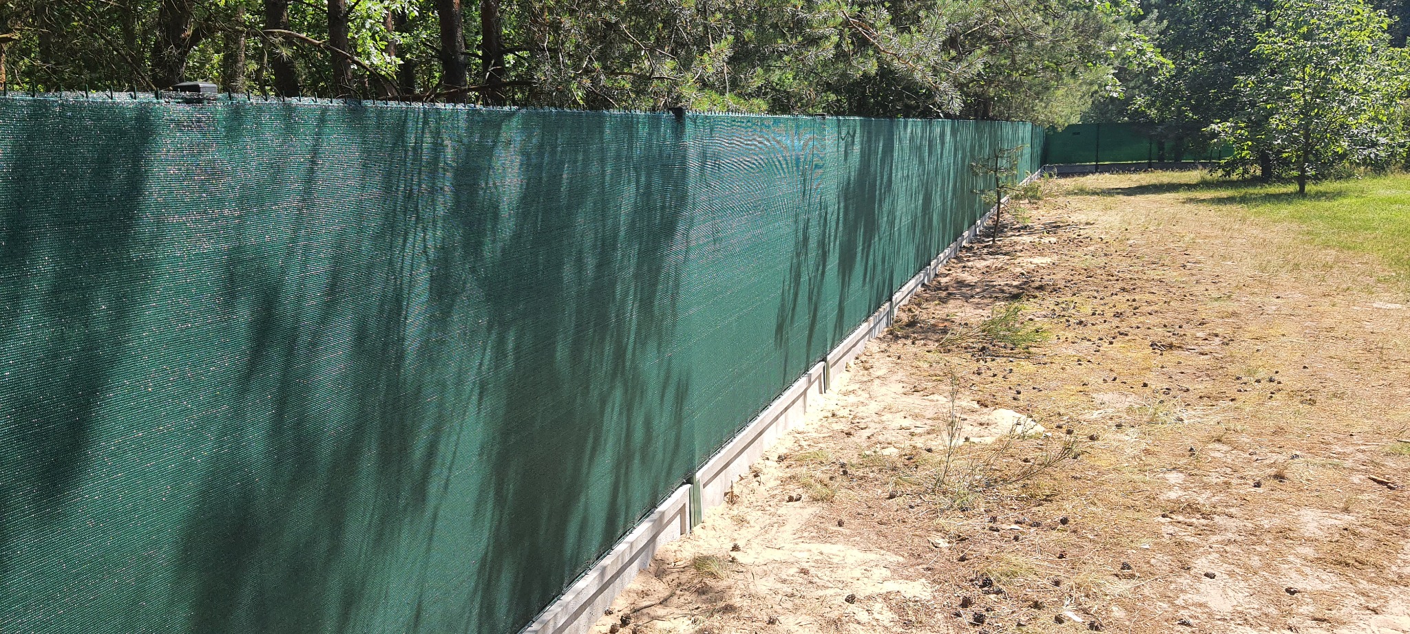 Długi, zielony płot z siatki maskującej na betonowej podmurówce, chroniący posesję przed wzrokiem przechodniów, z widocznymi cieniami drzew na powierzchni siatki.