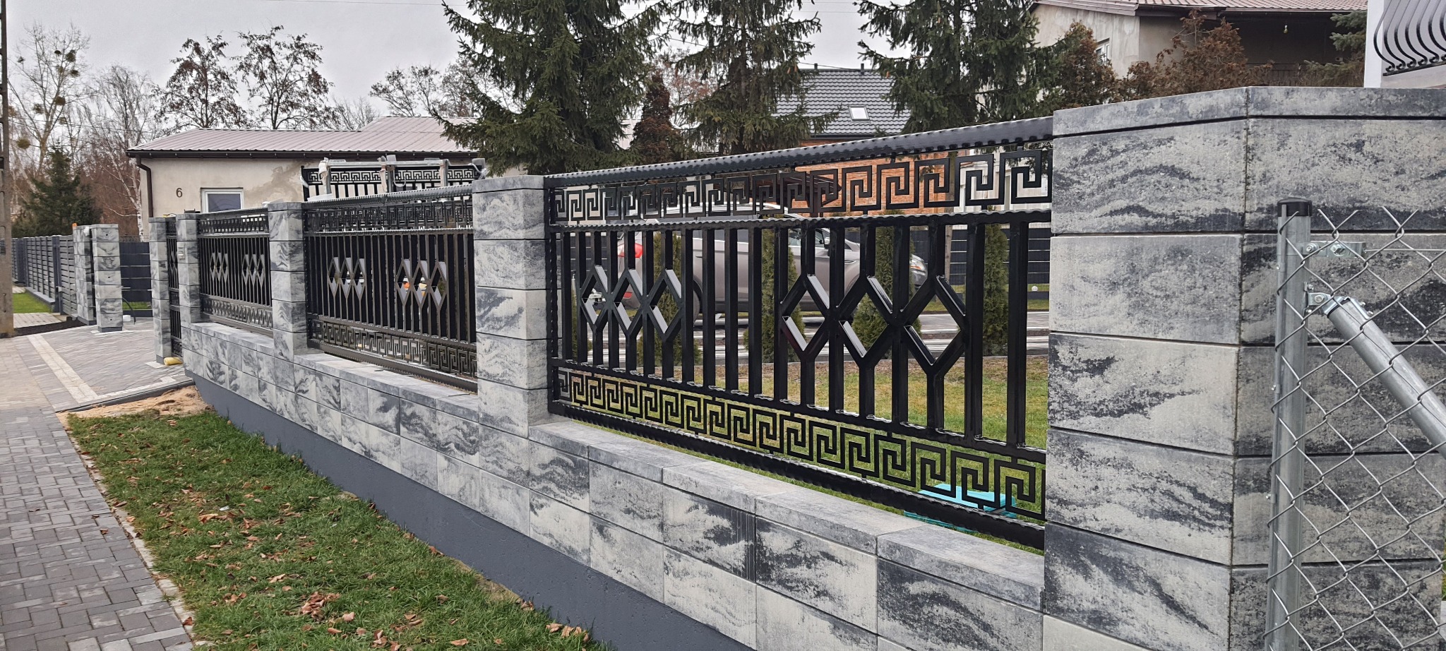 Nowoczesne ogrodzenie z czarnych metalowych przęseł z geometrycznym wzorem i słupków z szarego betonu architektonicznego, widok z boku na tle domów i drzew.