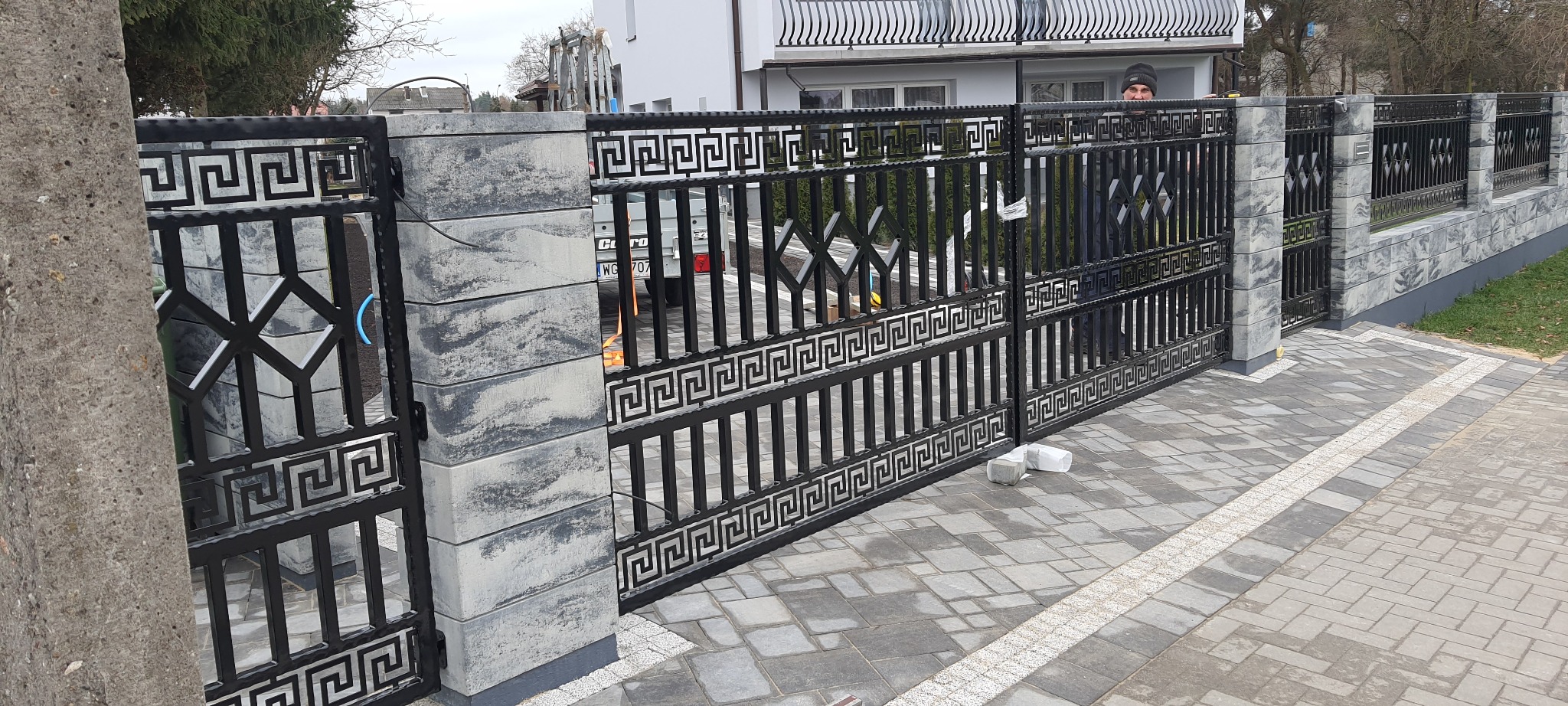 Czarna, metalowa brama przesuwna z motywem greckim, osadzona na betonowych słupkach z szarego, cieniowanego betonu, prowadząca na posesję z kostki brukowej; w tle widoczny fragment domu i pracownik...