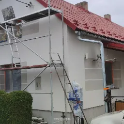 Dom jednorodzinny w trakcie malowania elewacji na biało, z widocznymi rusztowaniami, zabezpieczonymi oknami i częściowo pomalowaną fasadą