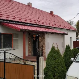 Dom jednorodzinny z czerwoną dachówką, częściowo ocieplony, z widocznym rusztowaniem, zabezpieczonym oknem oraz elementami dekoracyjnymi na elewacji, z samochodem zaparkowanym przed budynkiem.