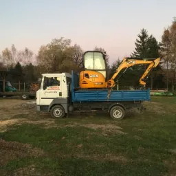 Mała koparka JCB na niebieskiej platformie transportowej, zaparkowana na trawiastym terenie z widocznym traktorem i budynkami w tle, o zmierzchu.