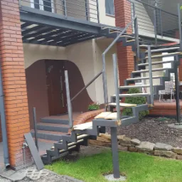 Zewnętrzne schody metalowe z drewnianymi stopniami prowadzące na taras z balustradą. Konstrukcja pomalowana na szaro, widoczna ceglana elewacja budynku.