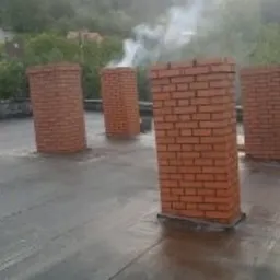 Praca na dachu: murarz w szarej bluzie w kapturze wznosi komin z czerwonej cegły. W tle zalesione wzgórza i mgła.