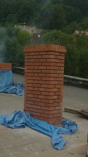 Murowany komin z cegły na dachu, zabezpieczony niebieską folią, widok z góry na tle zieleni.