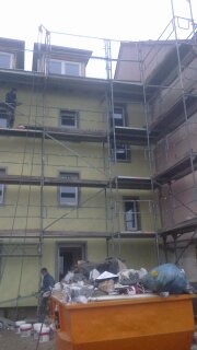 Budynek w trakcie malowania elewacji, otoczony rusztowaniami, z widocznym kontenerem na odpady i farby w wiadrach na pierwszym planie.