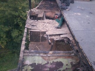 Demontaż fragmentu dachu z widocznymi uszkodzeniami, gruzem i odkrytą konstrukcją więźby dachowej. Pracownik w zielonych spodniach używa piły łańcuchowej. Widok z góry na częściowo rozebrany dach...