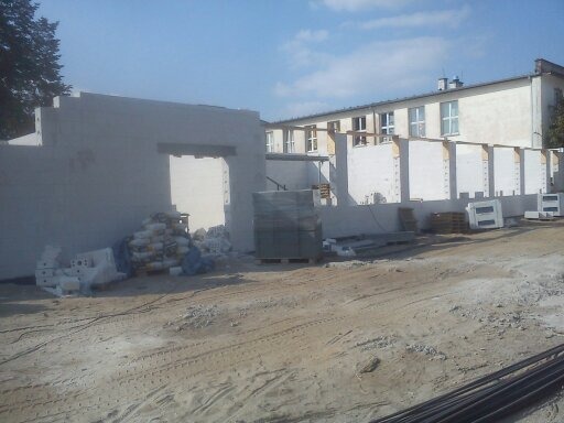 Budowa ściany budynku z prefabrykatów betonowych, widoczne worki z zaprawą, palety z materiałami budowlanymi i fragment budynku w tle