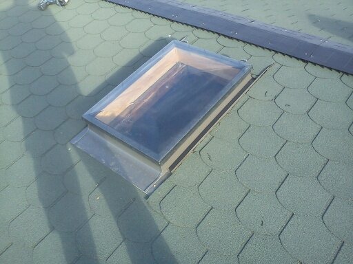 Zielony dach pokryty gontem bitumicznym z zamontowanym oknem dachowym z metalową obróbką blacharską, widoczny cień osoby wykonującej prace.