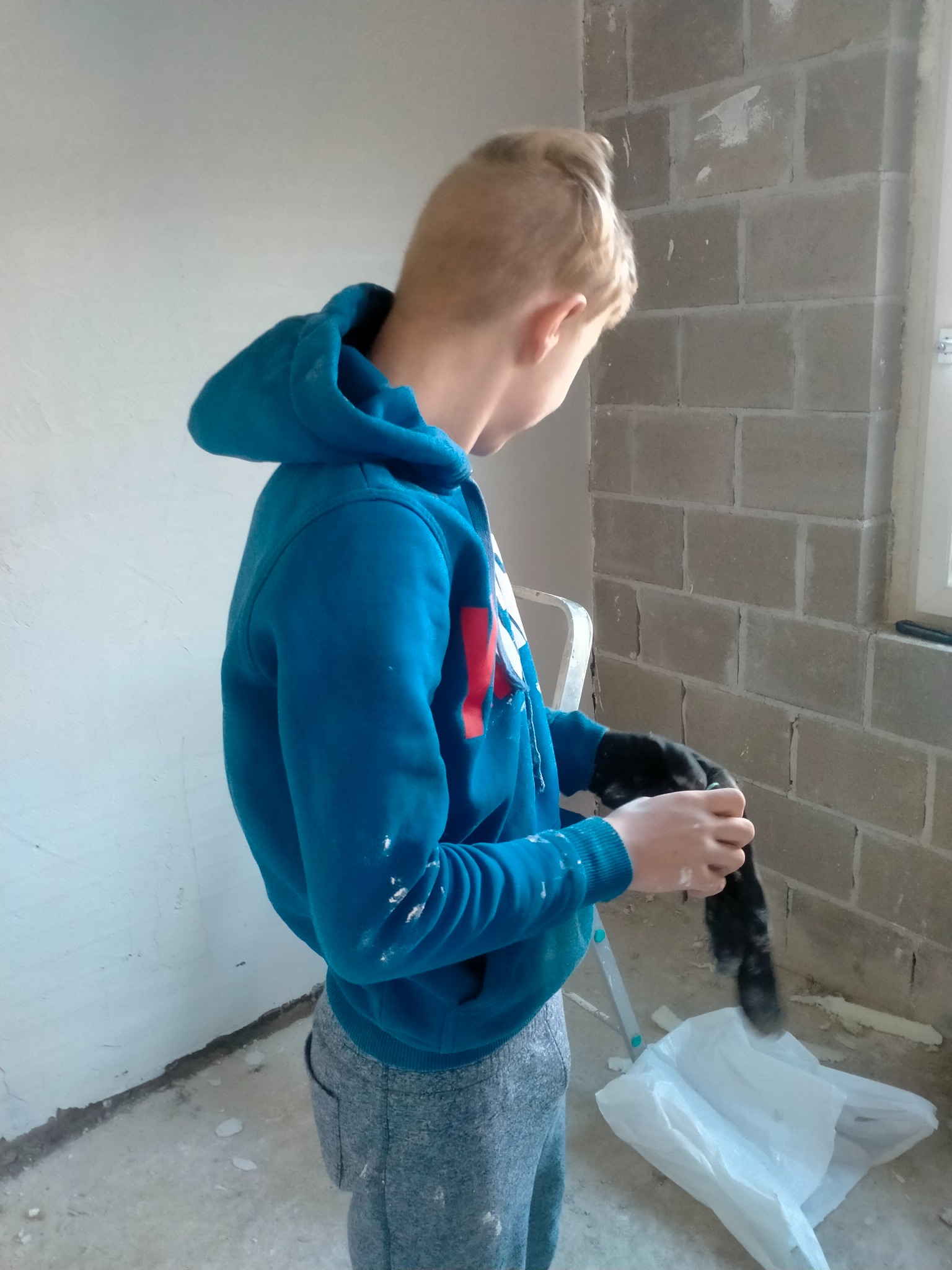 Młody mężczyzna w niebieskiej bluzie z kapturem czyści pędzel nad plastikowym workiem w trakcie prac wykończeniowych w surowym wnętrzu z ścianami z betonu komórkowego, na których widać ślady po...