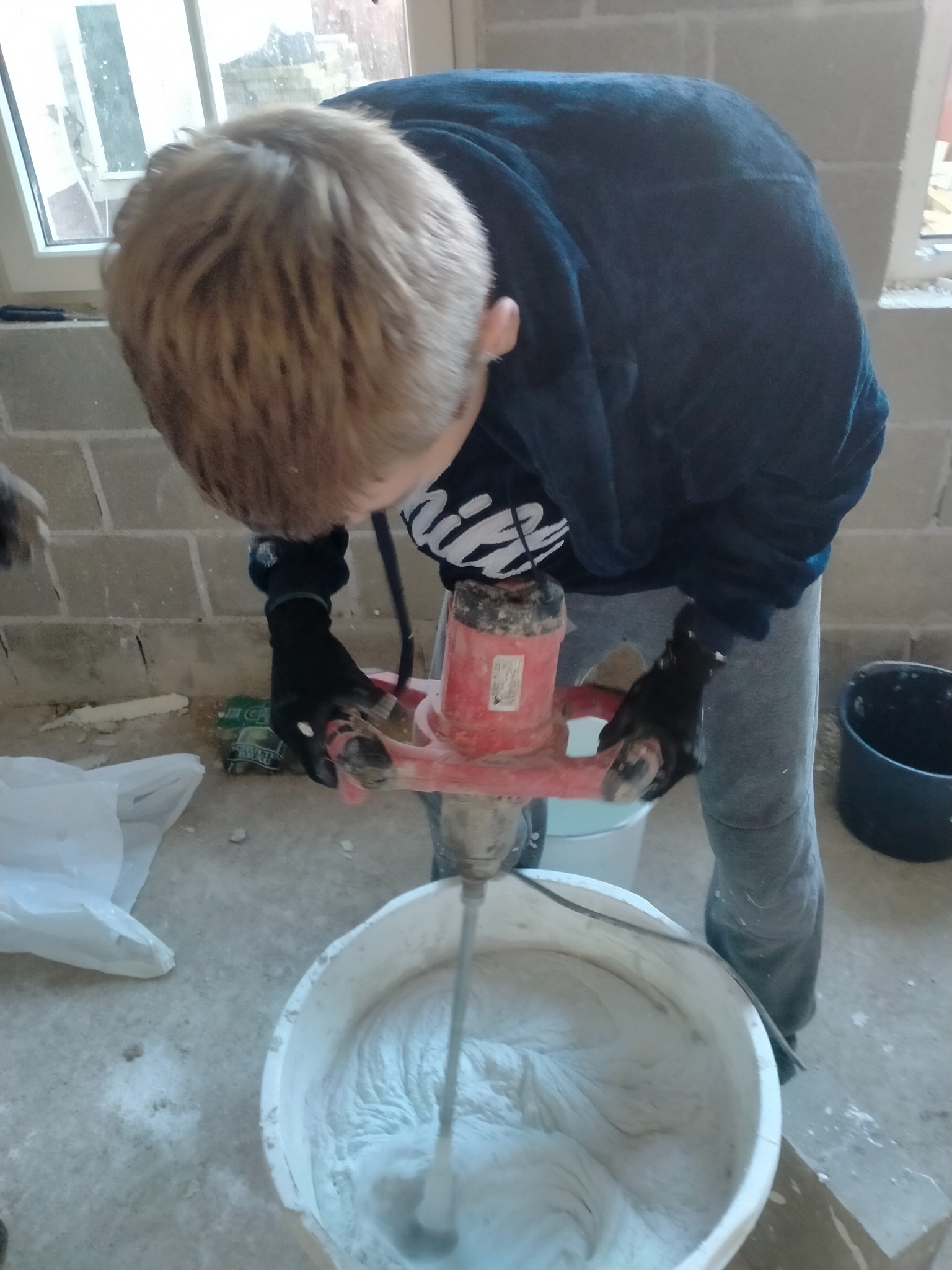 Mężczyzna w bluzie i rękawicach miesza zaprawę w wiadrze za pomocą mieszadła elektrycznego, widoczne ściany z bloczków betonowych w surowym stanie.