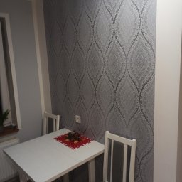 KAZDY SZCZEGÓŁ DOPRACOWANY - Pokój z szarą tapetą w ornamenty, biały stół z dekoracją świąteczną i dwa krzesła z szarymi siedziskami.