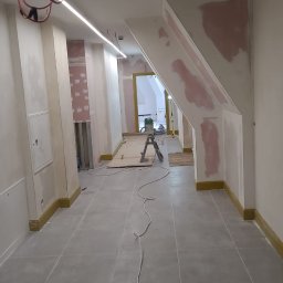 KAZDY SZCZEGÓŁ DOPRACOWANY - Korytarz w trakcie prac wykończeniowych: ściany z nałożoną gładzią szpachlową, widoczne przewody elektryczne pod sufitem, drabina rozstawiona w głębi pomieszczenia, szare płytki podłogowe.