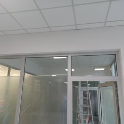 KAZDY SZCZEGÓŁ DOPRACOWANY - Biuro z nowym sufitem kasetonowym i przeszklonymi ścianami, zabezpieczonymi folią malarską, drabina oparta o ścianę.