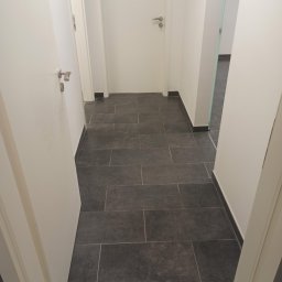 KAZDY SZCZEGÓŁ DOPRACOWANY - Korytarz z szarą, prostokątną podłogą z płytek, białe ściany i drzwi z srebrnymi klamkami, widok zza otwartych drzwi.