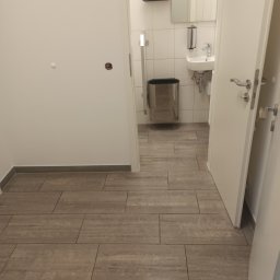 KAZDY SZCZEGÓŁ DOPRACOWANY - Wykończona łazienka z szarymi płytkami podłogowymi imitującymi drewno, widok z korytarza przez otwarte drzwi na umywalkę i kosz na śmieci.