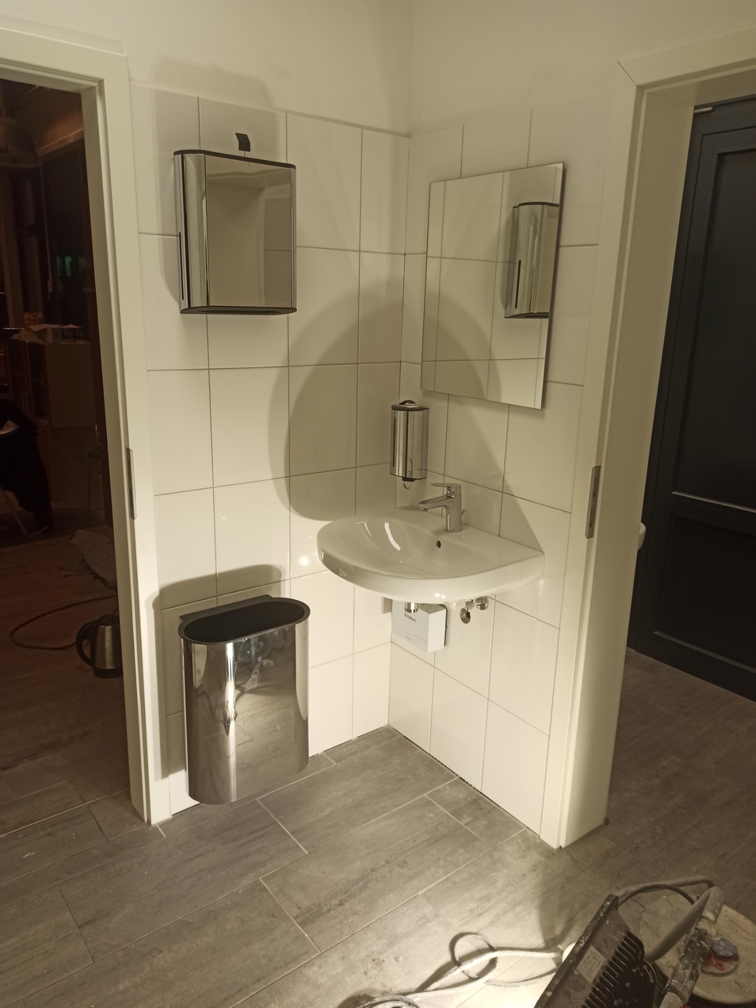 Niewielka łazienka z białą umywalką narożną, lustrem, podajnikiem na mydło i stalowym koszem na śmieci, wykończona białymi płytkami i szarą podłogą z płytek imitujących drewno, widoczny sprzęt...