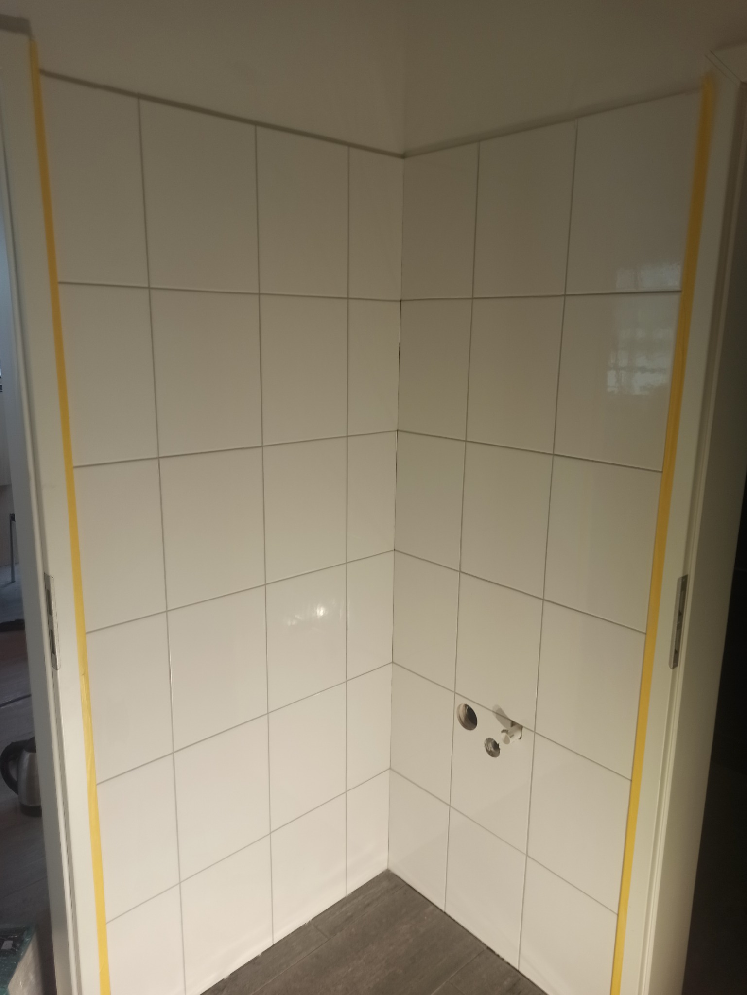 Świeżo wykafelkowana narożna ściana kabiny prysznicowej z widocznymi otworami na baterię, zabezpieczona żółtą taśmą malarską na framugach drzwi.