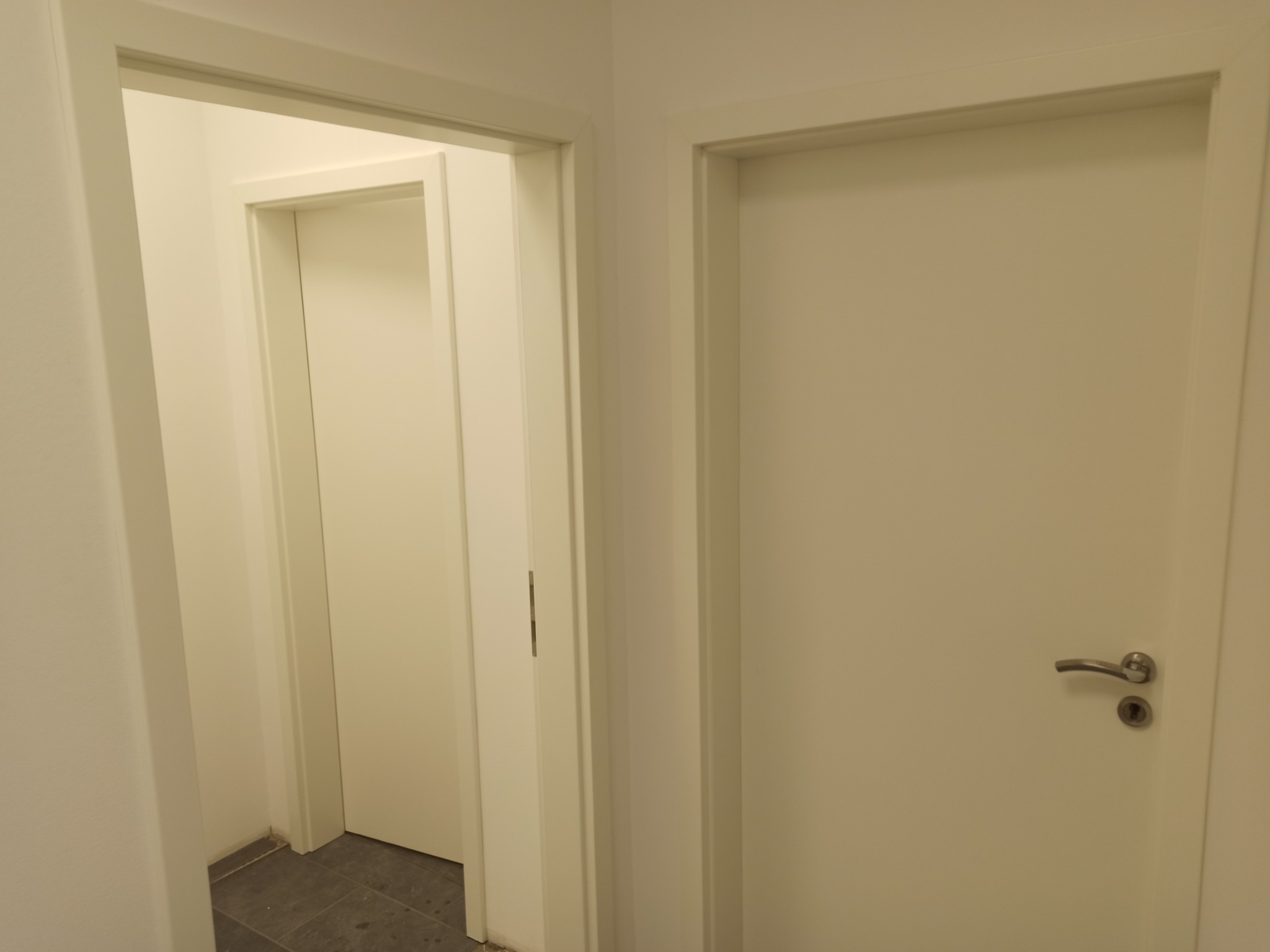 Dwa białe, pomalowane drzwi wewnętrzne w perspektywie, widoczna klamka na jednych z nich, prowadzące do kolejnego pomieszczenia z szarą podłogą.