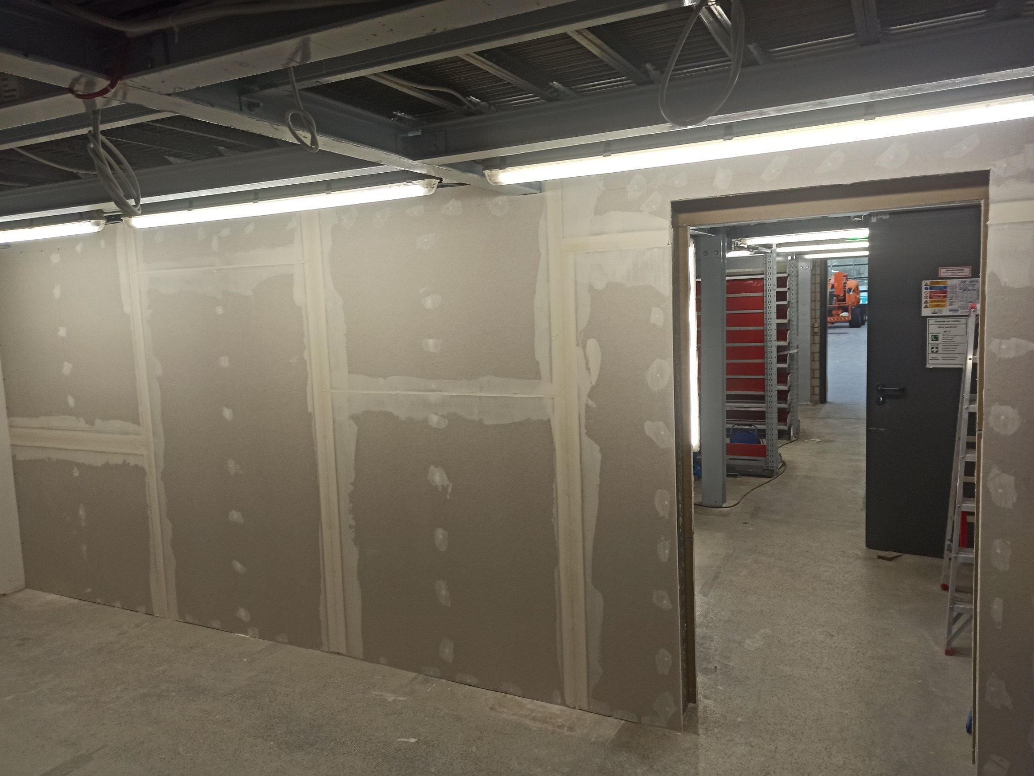 Montaż płyt kartonowo-gipsowych w pomieszczeniu biurowym, widoczne łączenia szpachlowane, sufit z instalacjami, otwarte drzwi do dalszej części budynku.