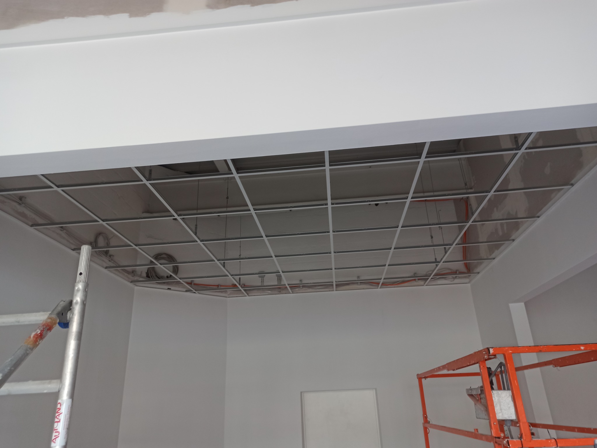 Montaż sufitu podwieszanego z widoczną konstrukcją metalową, instalacją elektryczną i fragmentem drabiny oraz podnośnika.