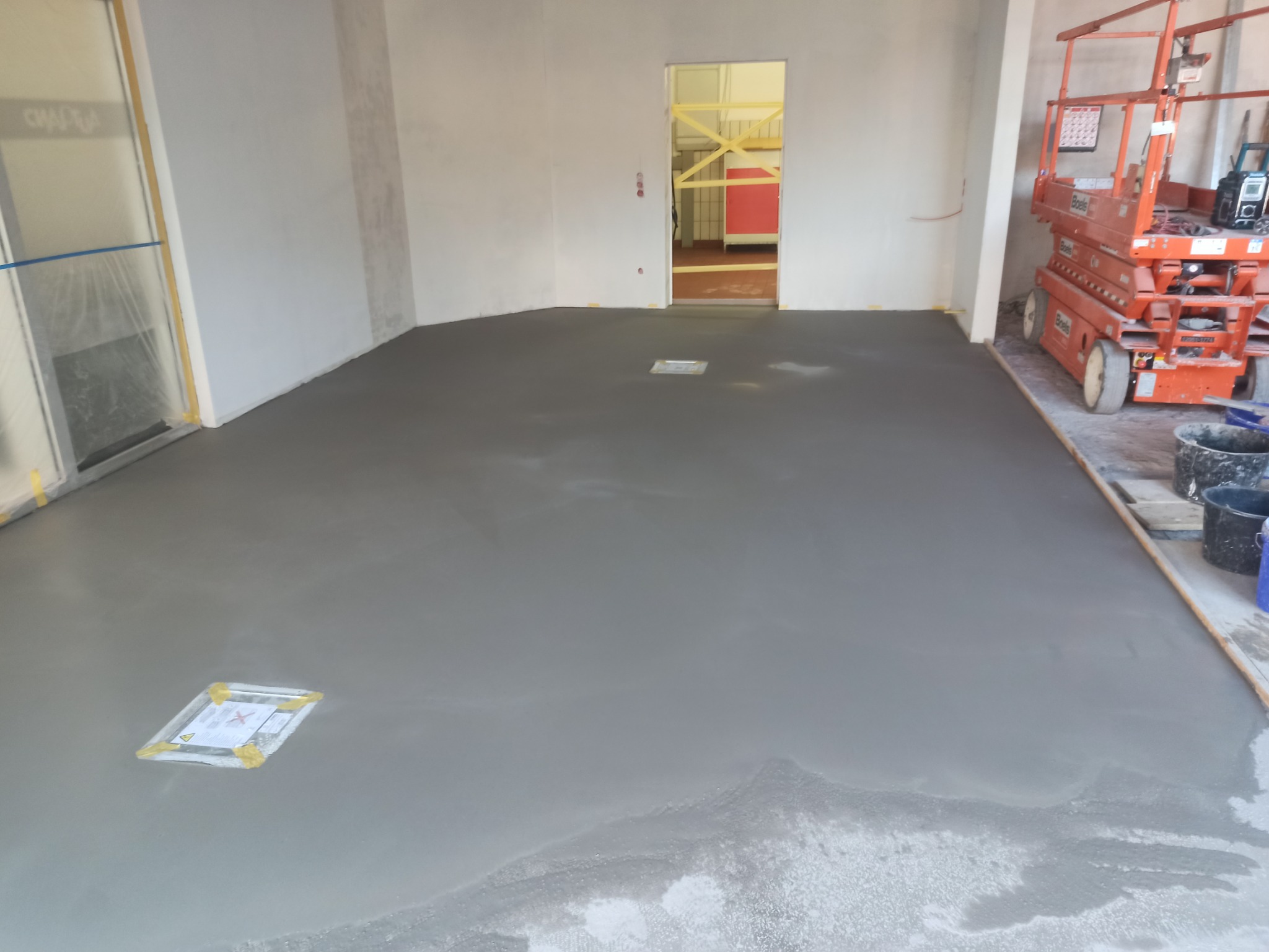 Wylewka samopoziomująca na podłodze w trakcie wysychania, zabezpieczona folią i taśmą kratka wentylacyjna, w tle podnośnik nożycowy.