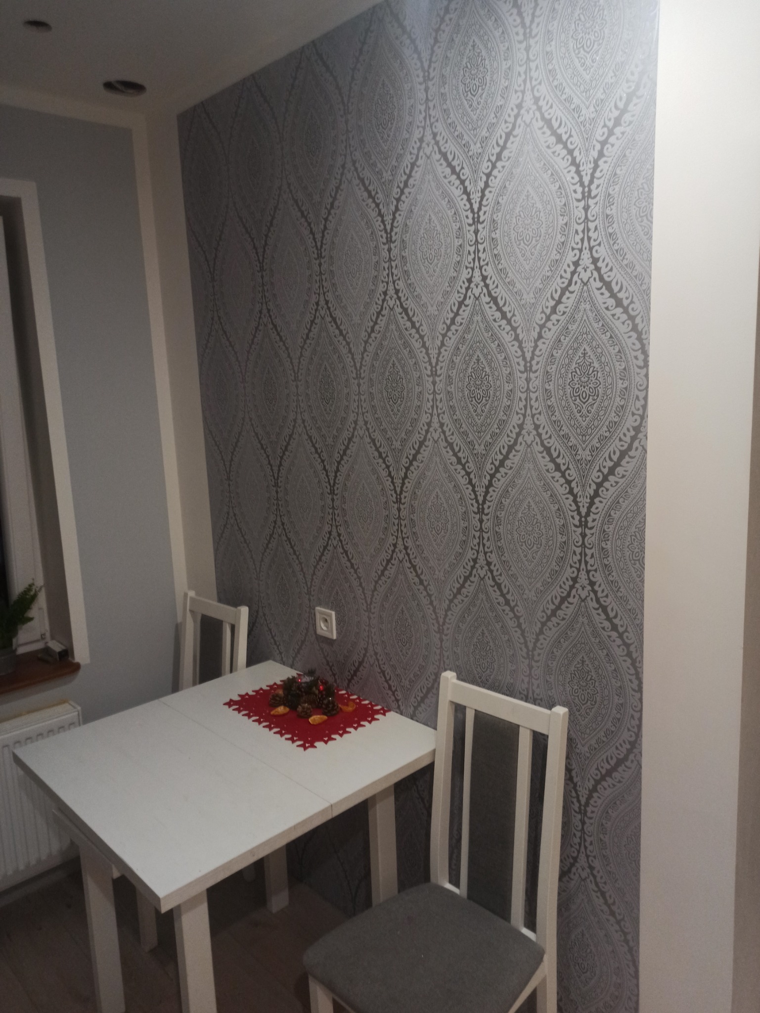 Pokój z szarą tapetą w ornamenty, biały stół z dekoracją świąteczną i dwa krzesła z szarymi siedziskami.