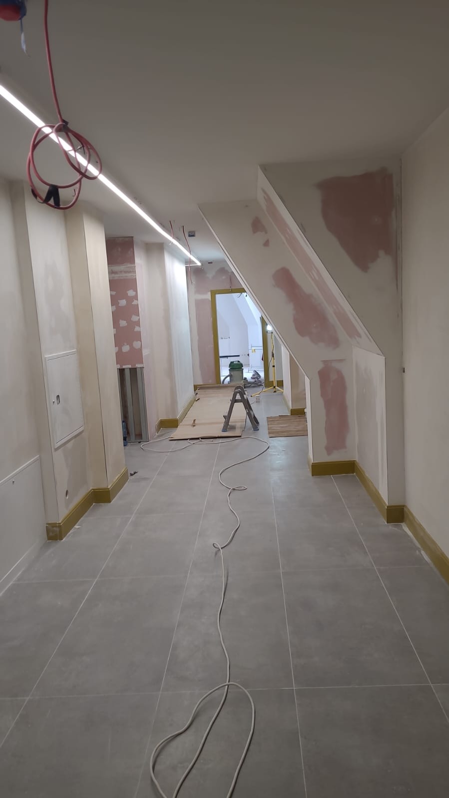 Korytarz w trakcie prac wykończeniowych: ściany z nałożoną gładzią szpachlową, widoczne przewody elektryczne pod sufitem, drabina rozstawiona w głębi pomieszczenia, szare płytki podłogowe.