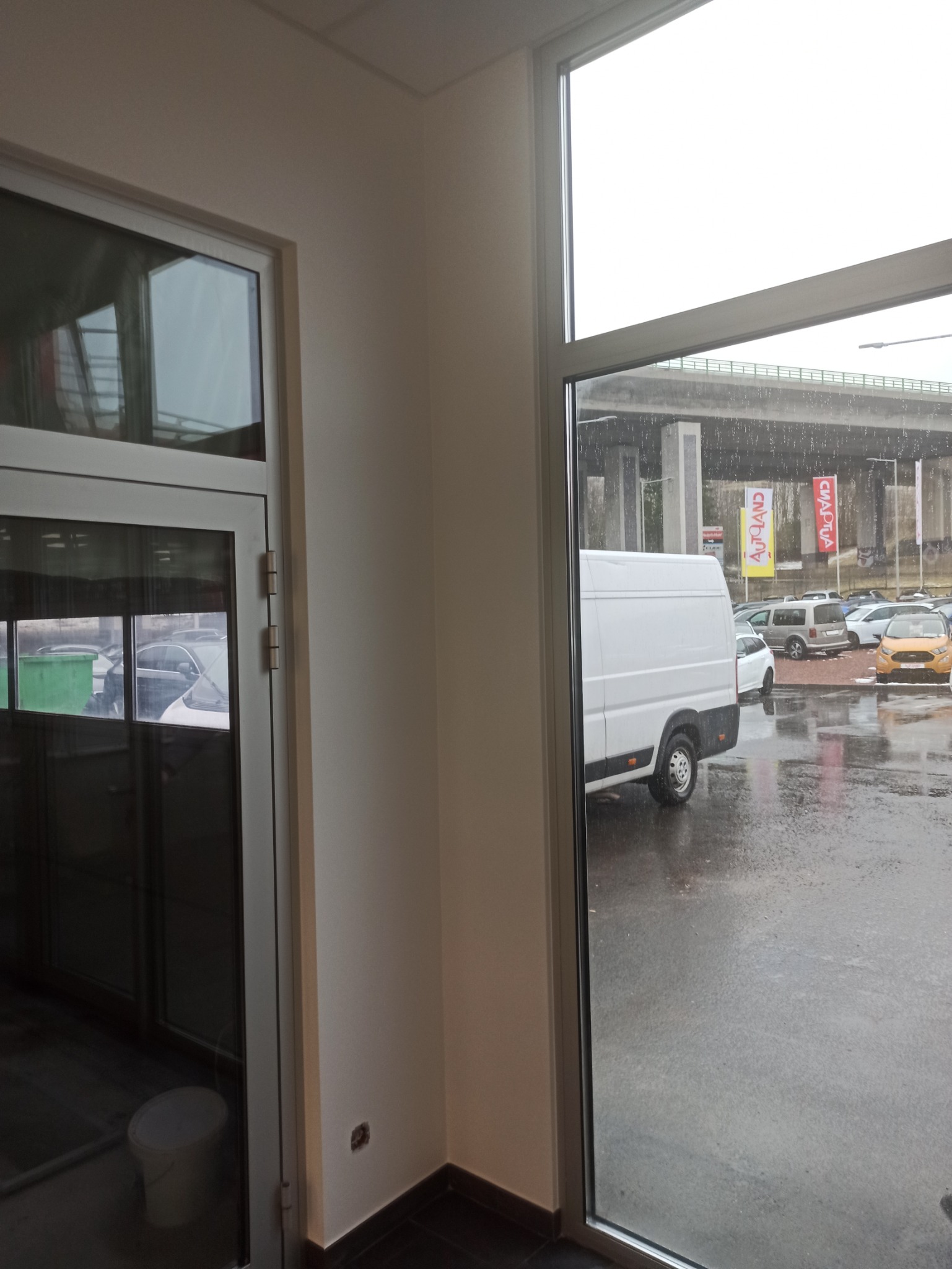 Wnętrze biura z widokiem na deszczowy parking i białą furgonetkę przez duże okno.