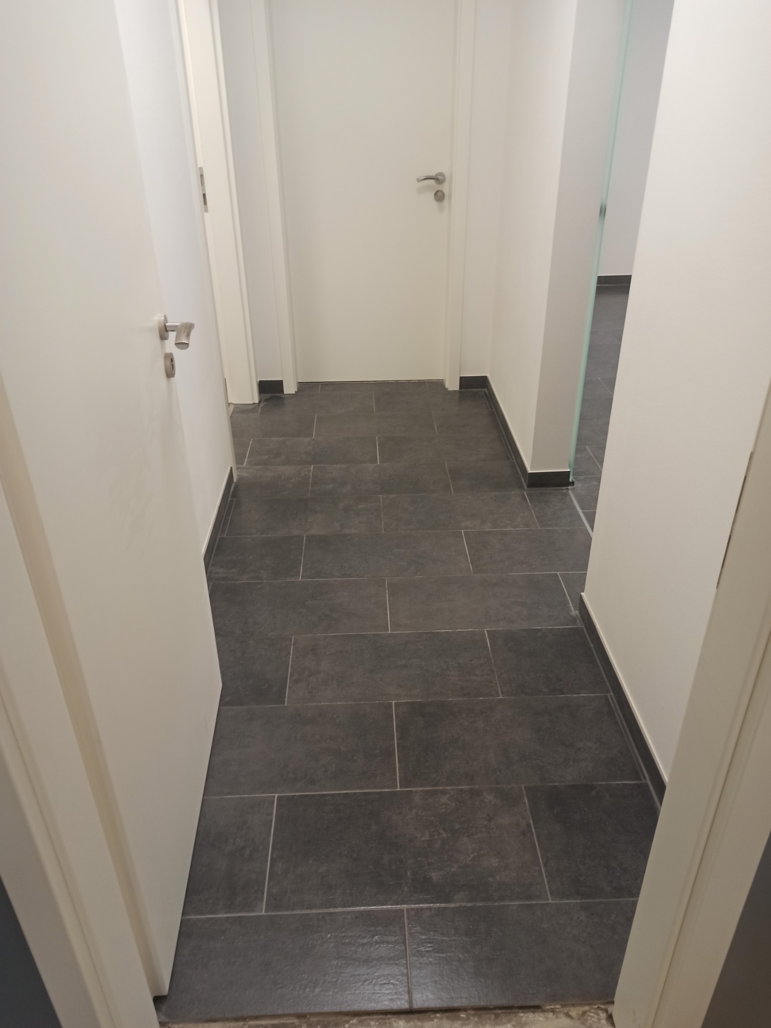 Korytarz z szarą, prostokątną podłogą z płytek, białe ściany i drzwi z srebrnymi klamkami, widok zza otwartych drzwi.