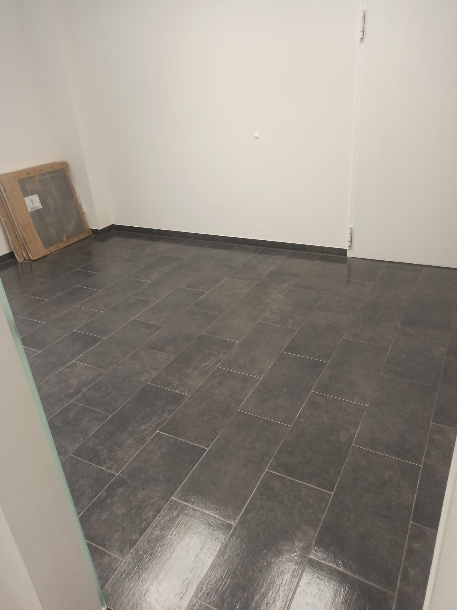 Ułożona ciemnoszara podłoga z płytek w pustym pomieszczeniu z białymi ścianami i drzwiami, widoczne paczki płytek w rogu.