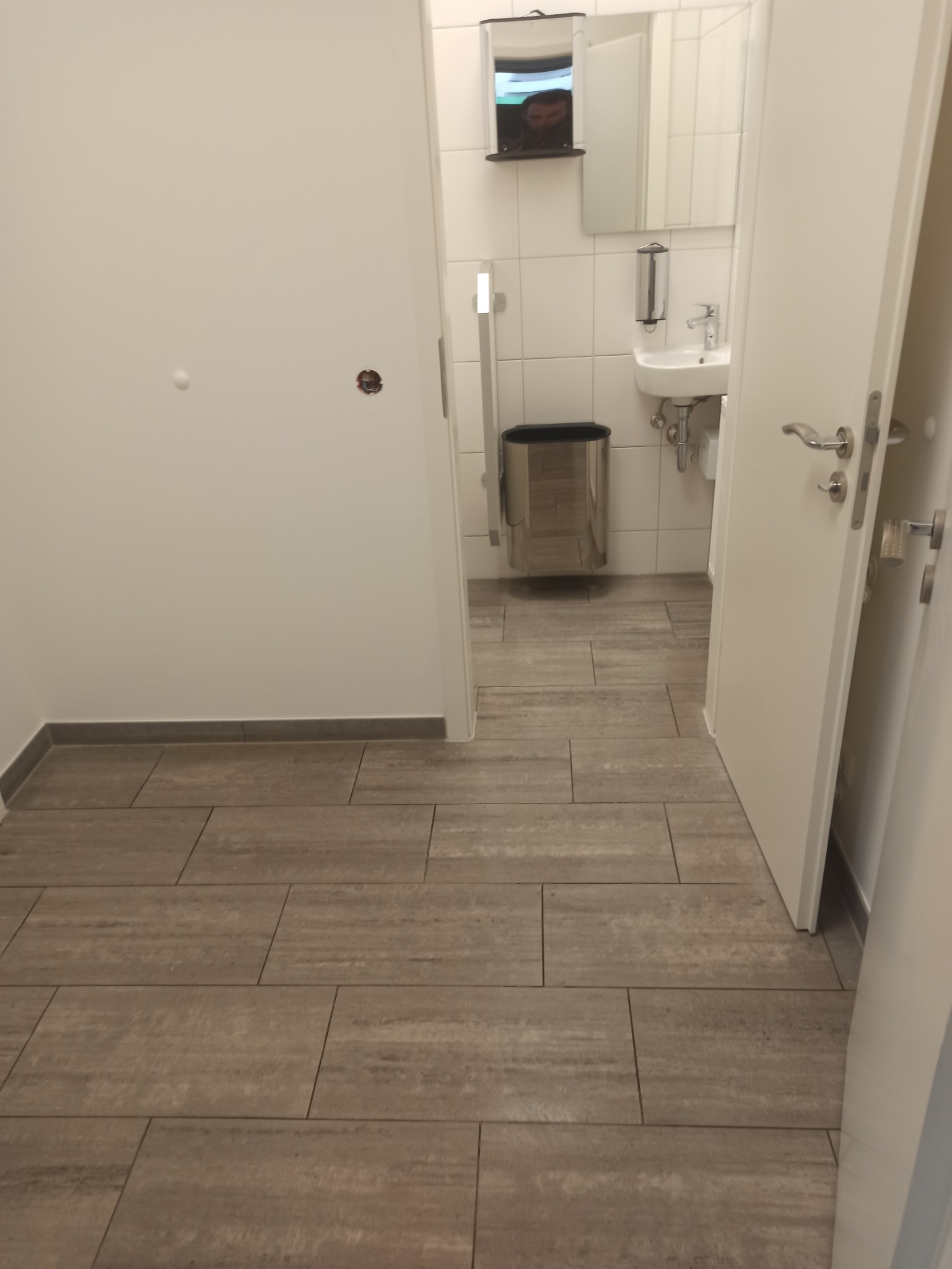 Wykończona łazienka z szarymi płytkami podłogowymi imitującymi drewno, widok z korytarza przez otwarte drzwi na umywalkę i kosz na śmieci.