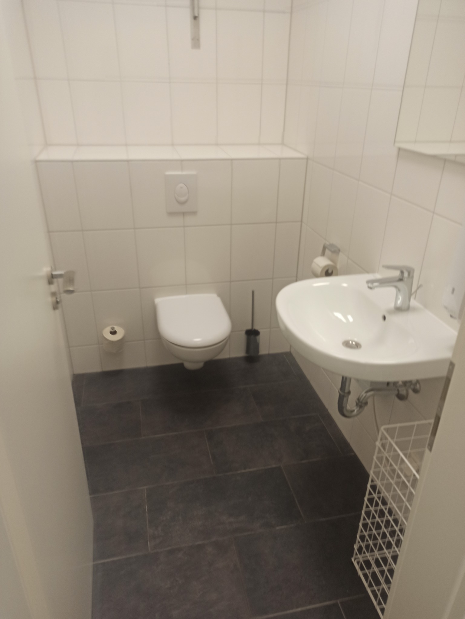 Nowoczesna łazienka z białymi płytkami, wiszącą umywalką i toaletą, uzupełniona o ciemne płytki podłogowe i minimalistyczne akcesoria.