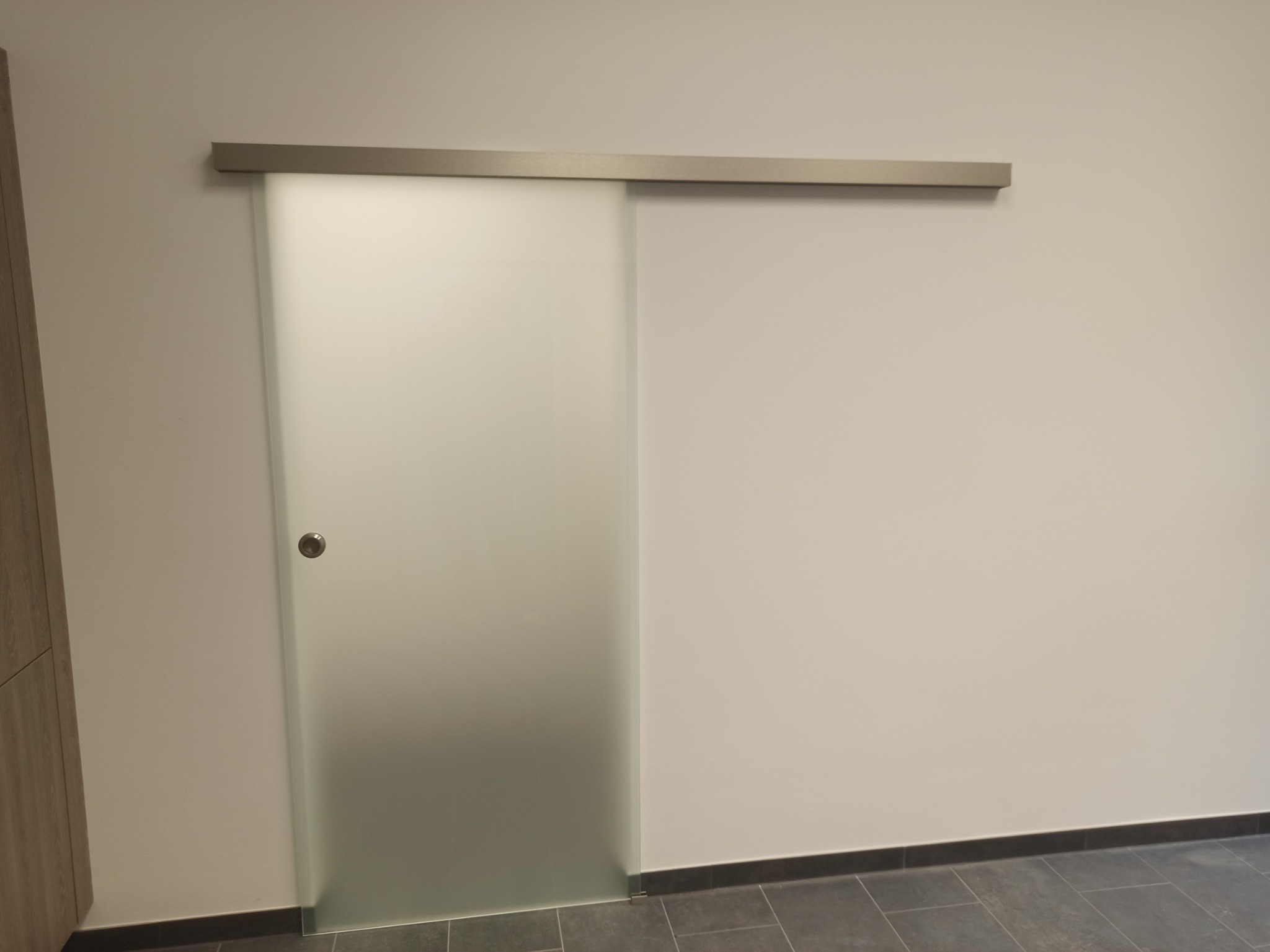 Szklane drzwi przesuwne z matową powierzchnią i metalową prowadnicą, zamontowane w jasnym pomieszczeniu z szarą podłogą.