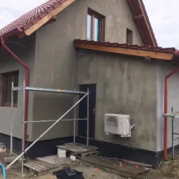 Dom w trakcie nakładania szarego tynku elewacyjnego, widoczne rusztowania, zamontowany klimatyzator na ścianie, czerwone rynny i dachówka.