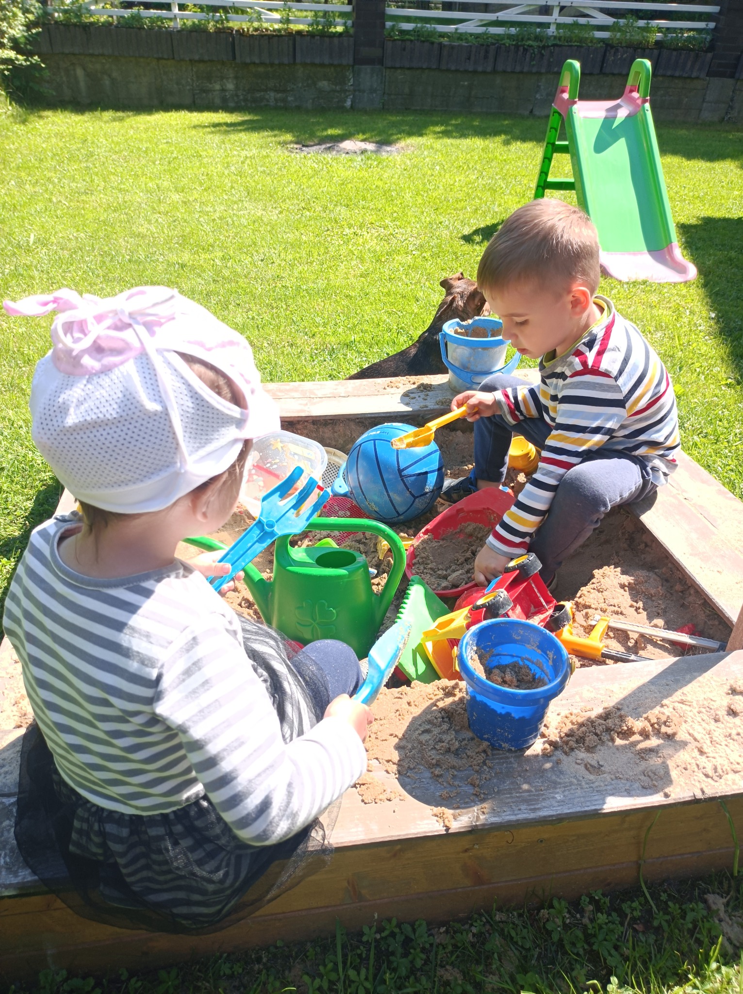 Dwoje dzieci w paski bawi się w piaskownicy pełnej zabawek na tle trawnika i zjeżdżalni.