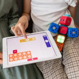 Dziewczynka w jasnej koszulce i kraciastej spódnicy siedzi na zielonej kanapie i bawi się interaktywnymi kostkami edukacyjnymi, programując je za pomocą tabletu z interfejsem graficznym.