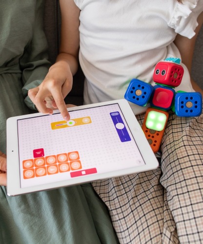 Dziewczynka w jasnej koszulce i kraciastej spódnicy siedzi na zielonej kanapie i bawi się interaktywnymi kostkami edukacyjnymi, programując je za pomocą tabletu z interfejsem graficznym.