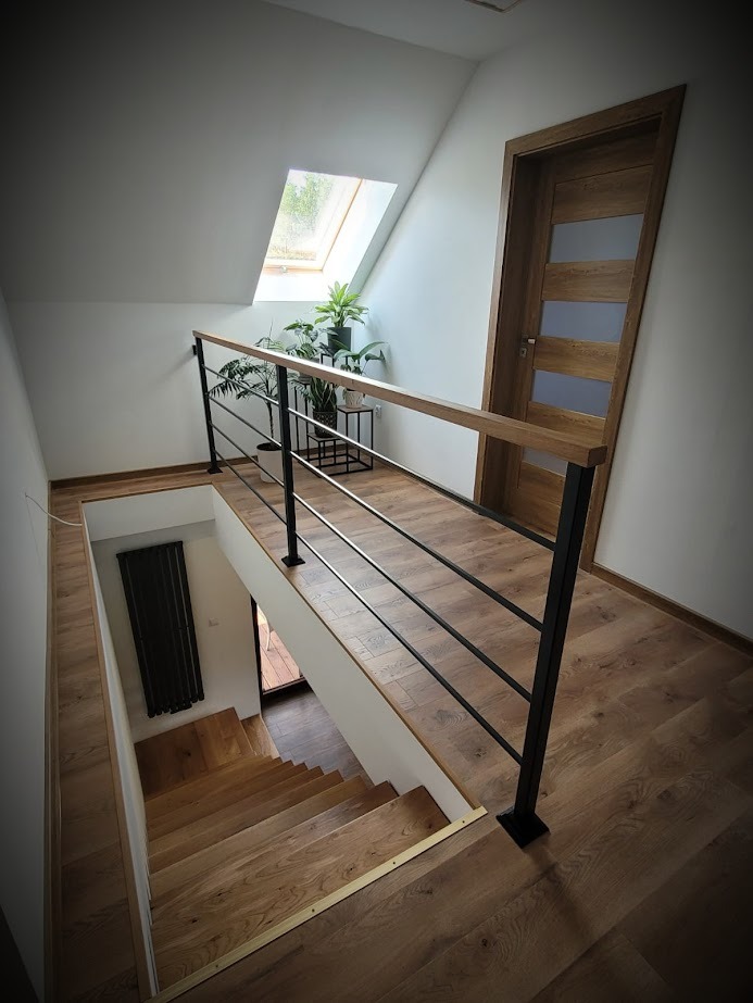 Nowoczesne poręcze schodowe z czarnymi, poziomymi prętami i drewnianym wykończeniem, widok z góry na drewniane schody, okno dachowe i rośliny w tle.