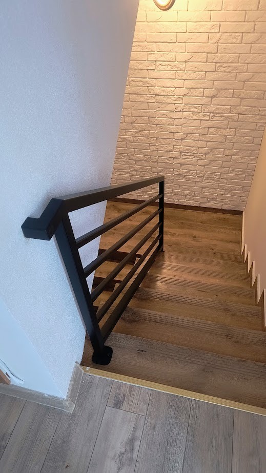 Nowoczesna, czarna poręcz schodowa z poziomymi prętami, zamontowana przy ścianie z dekoracyjną cegłą i drewnianymi schodami.